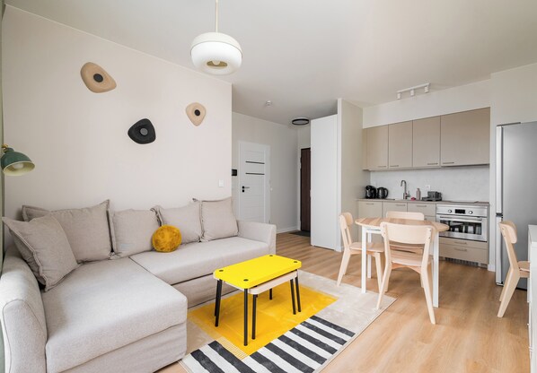 Ein zeitgemäßer Wohnraum mit Sofa, gelbem Tisch, Essbereich mit Stühlen sowie Küche mit Schränken und Geräten.