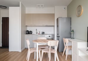 Moderni keittiö, jossa on valkoiset kaapit, ruostumattomasta teräksestä valmistettu jääkaappi, puinen ruokapöytä tuoleineen sekä pieni työtaso, jossa on lamppu.