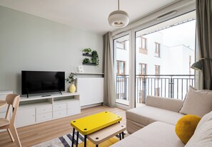 Moderni olohuone, jossa on litteänäyttöinen televisio, sohva, keltainen pöytä ja parveke, jolta on näkymä rakennuksiin.