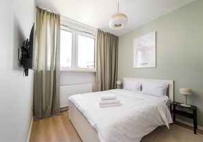 Ein Hotelzimmer mit Bett, Heizkörper, einem mit Vorhängen versehenen Fenster, einem an der Wand befestigten Fernseher, einer Nachttischlampe und einer gerahmten Karte an der Wand.