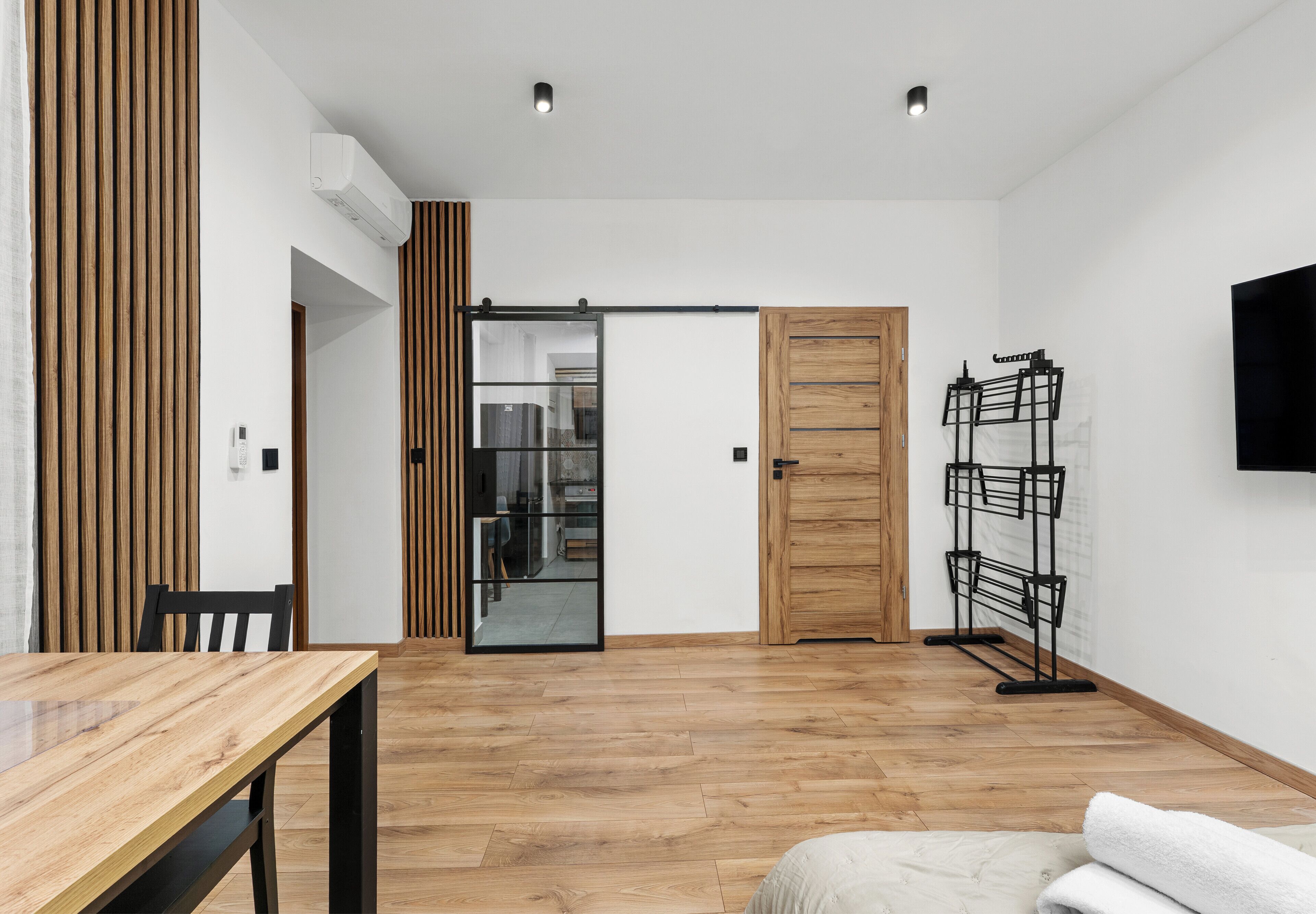 A modern room with a wooden floor, a sliding glass door, a wooden door, a TV mounted on the wall, and a black metal rack.