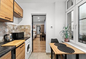 Eine kompakte Küche mit Mikrowelle, Wasserkocher und Waschmaschine. Es gibt einen Essbereich mit Tisch und Stühlen sowie einen Blumentopf am Fenster.