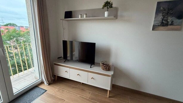 Ein modernes Wohnzimmer mit Fernseher auf einem weißen Schrank, ein Balkon mit Blick und einem Bild an der Wand.