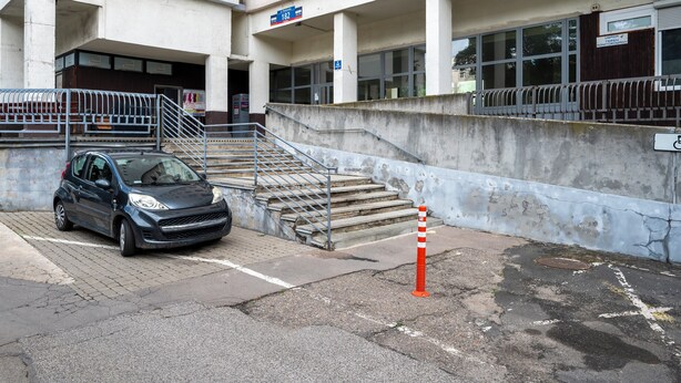 Um edifício com escadas que levam à entrada, um carro estacionado e o sinal de acessibilidade para cadeiras de rodas.