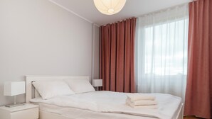 Uma cama bem arrumada com lençóis brancos, uma mesa de cabeceira com abajur e uma janela com cortinas leves e um tecido liso.