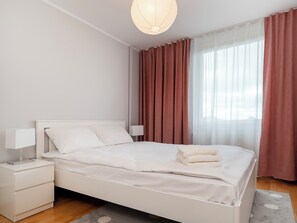A neatly made bed with white linens, a bedside table with a lamp, and a window with sheer curtains and a solid-colored drape. - Piotrkowska Spacious Apartment (Łódź)
