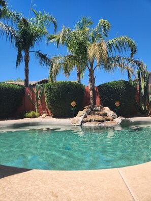 A clear pool surrounded by palm trees and cacti, with a rock feature in the center.