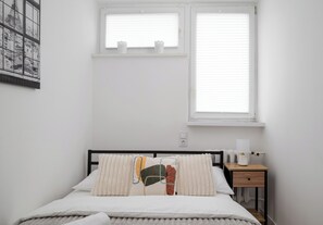 Un dormitorio con cama, mesita de noche, ventana y lámpara de pared.