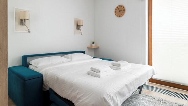Cama bem arrumada com lençóis e toalhas brancas, cabeceira azul, relógio de parede e uma pequena prateleira com uma planta.
