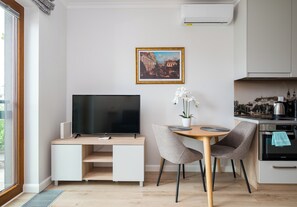 Een moderne keuken met een ingebouwde oven, een eettafel met stoelen, een televisie op een witte kast en een stadsgezicht in een lijst aan de muur.