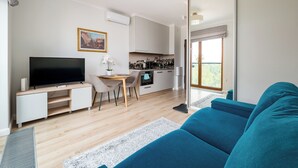 Uma sala de estar moderna com televisão de tela plana, um sofá, área de jantar com mesa e cadeiras, e cozinha ao fundo.