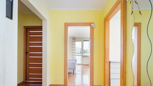 A hallway with wooden flooring, a door with horizontal slats, and a view into a room with a chair and a window. - Wilanów Comfort Stay Apartment (Warszawa)