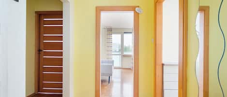 Un pasillo con piso de madera, una puerta con listones horizontales y vista a una habitación con una silla y un ventanal.