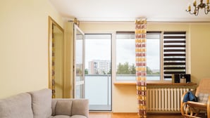 A living room with a sofa, a radiator, a window with blinds, and a view of buildings outside. - Wilanów Comfort Stay Apartment (Warszawa)