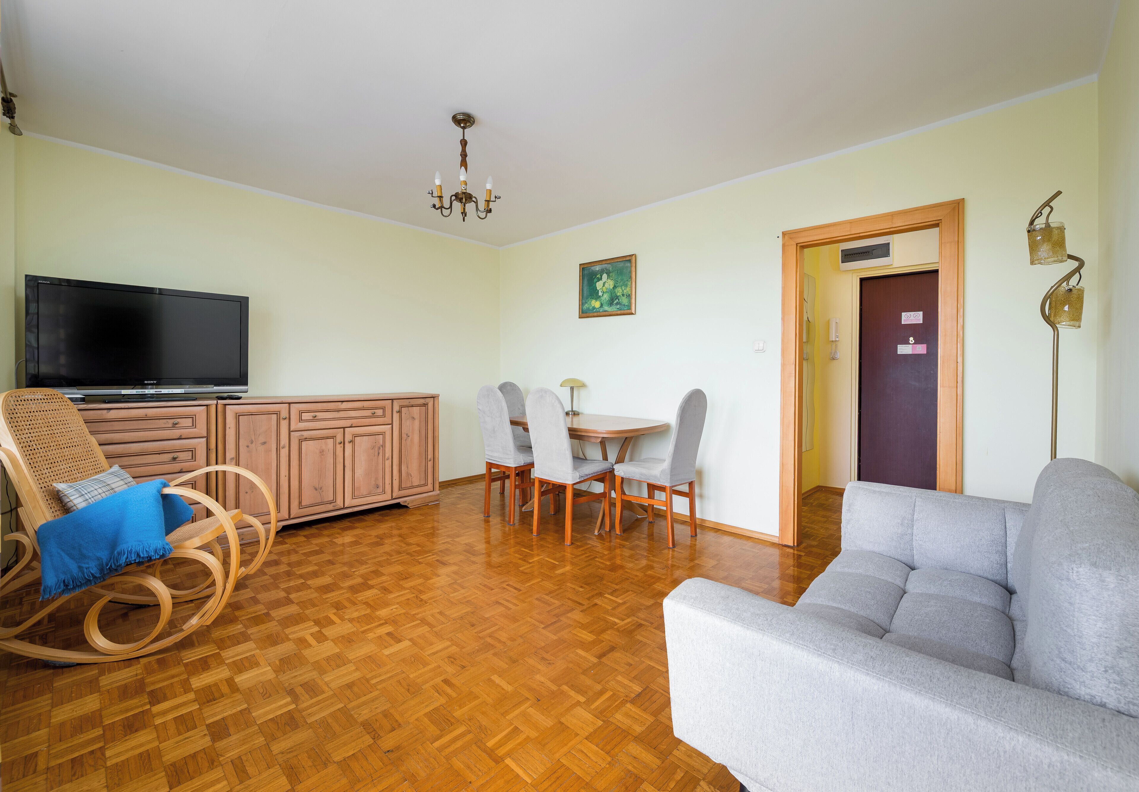 Een woonkamer met een bank, een televisie, een eettafel met stoelen, een houten kast en een kroonluchter.