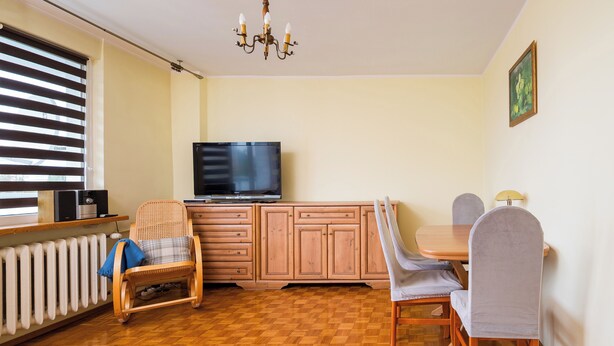 A living room with a television, wooden cabinets, a dining table with chairs, a radiator, and a window with blinds. - Wilanów Comfort Stay Apartment (Warszawa)
