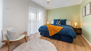 Ett sovrum med en säng, en stol, ett sängbord, en lampa, en takfläkt och ett fönster med tunna, genomskinliga gardiner.