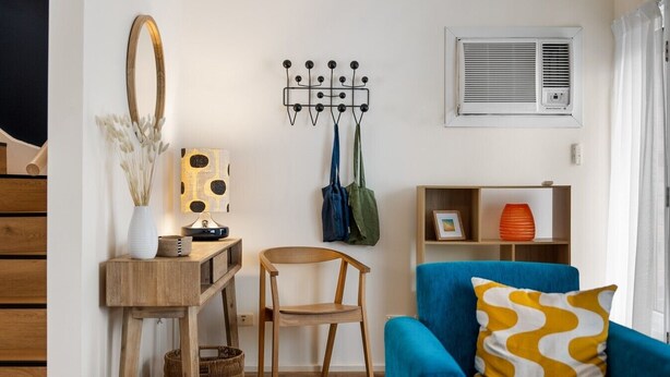A room with a wooden desk, a blue armchair with a patterned cushion, a wall-mounted mirror, a coat rack, and a shelf with decorative items.