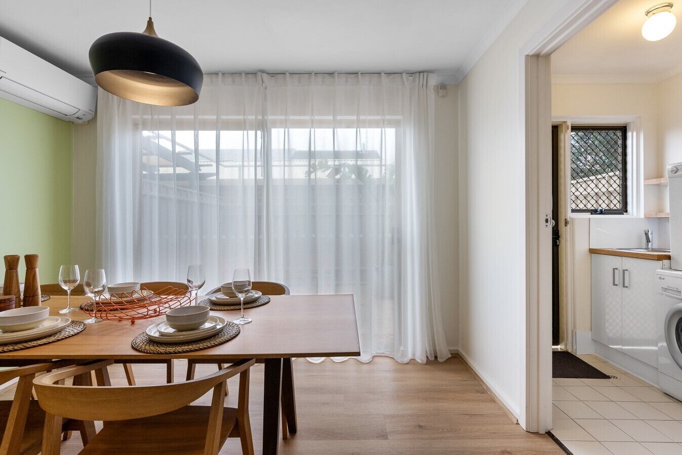 Un coin repas avec une table dressée pour quatre personnes, une fenêtre ornée de rideaux légers et une vue sur un bâtiment à l’extérieur.