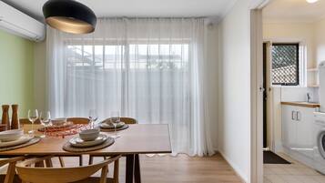 Un coin repas avec une table dressée pour quatre personnes, une fenêtre ornée de rideaux légers et une vue sur un bâtiment à l’extérieur.