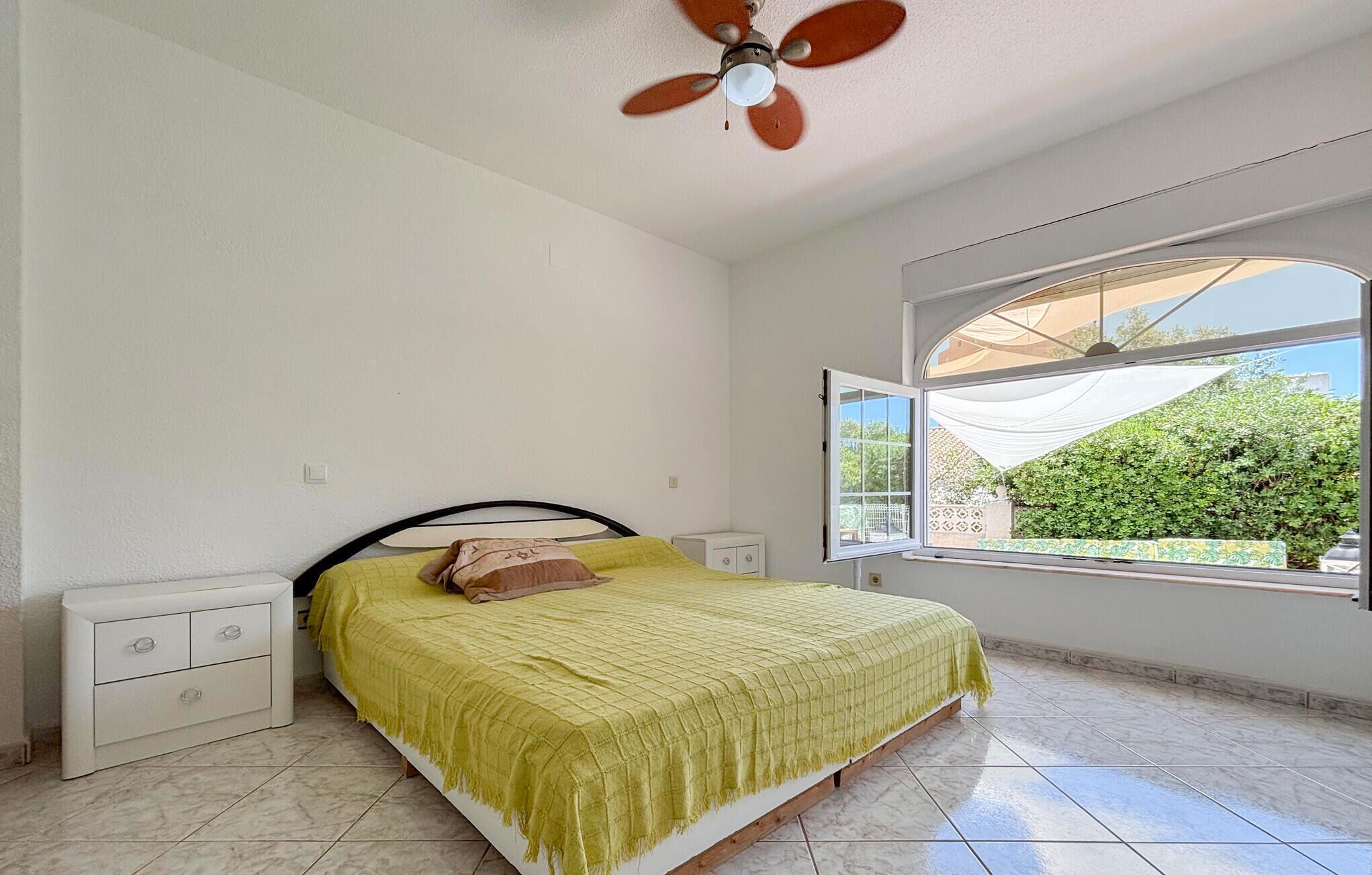A bedroom with a bed, a nightstand, a ceiling fan, a window with a view of greenery, and a tiled floor.