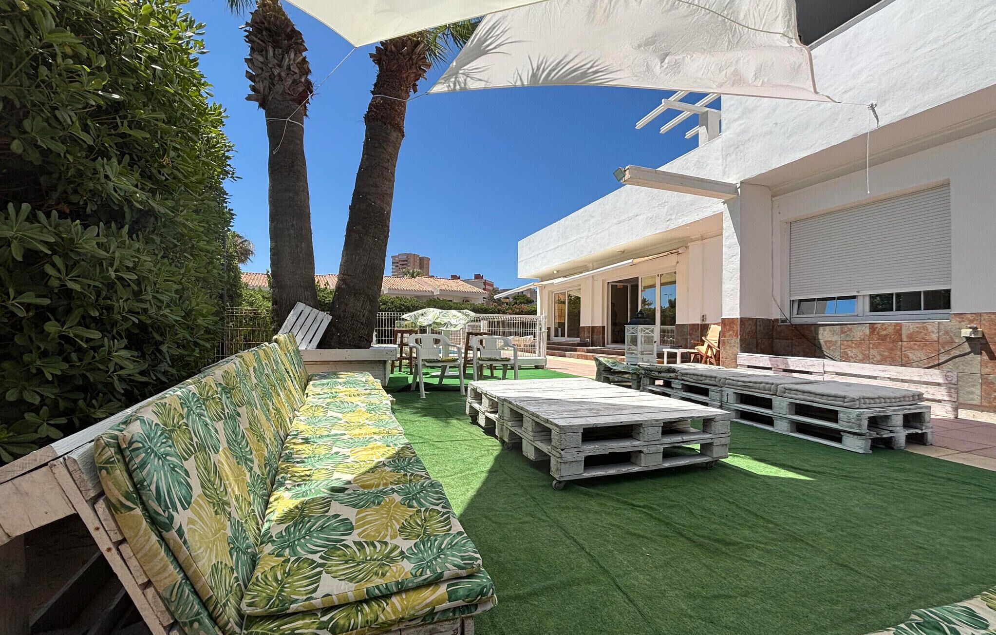 An outdoor seating area with a sofa, tables, and chairs, surrounded by greenery and palm trees.