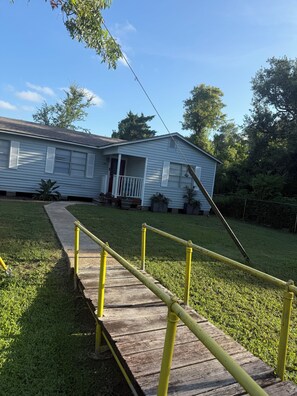 A residential property with a wooden deck, a yellow safety railing, and a house with white siding and a red door. - Mermaid by the Coast  4BR Near Beach & Refineries (Clute)