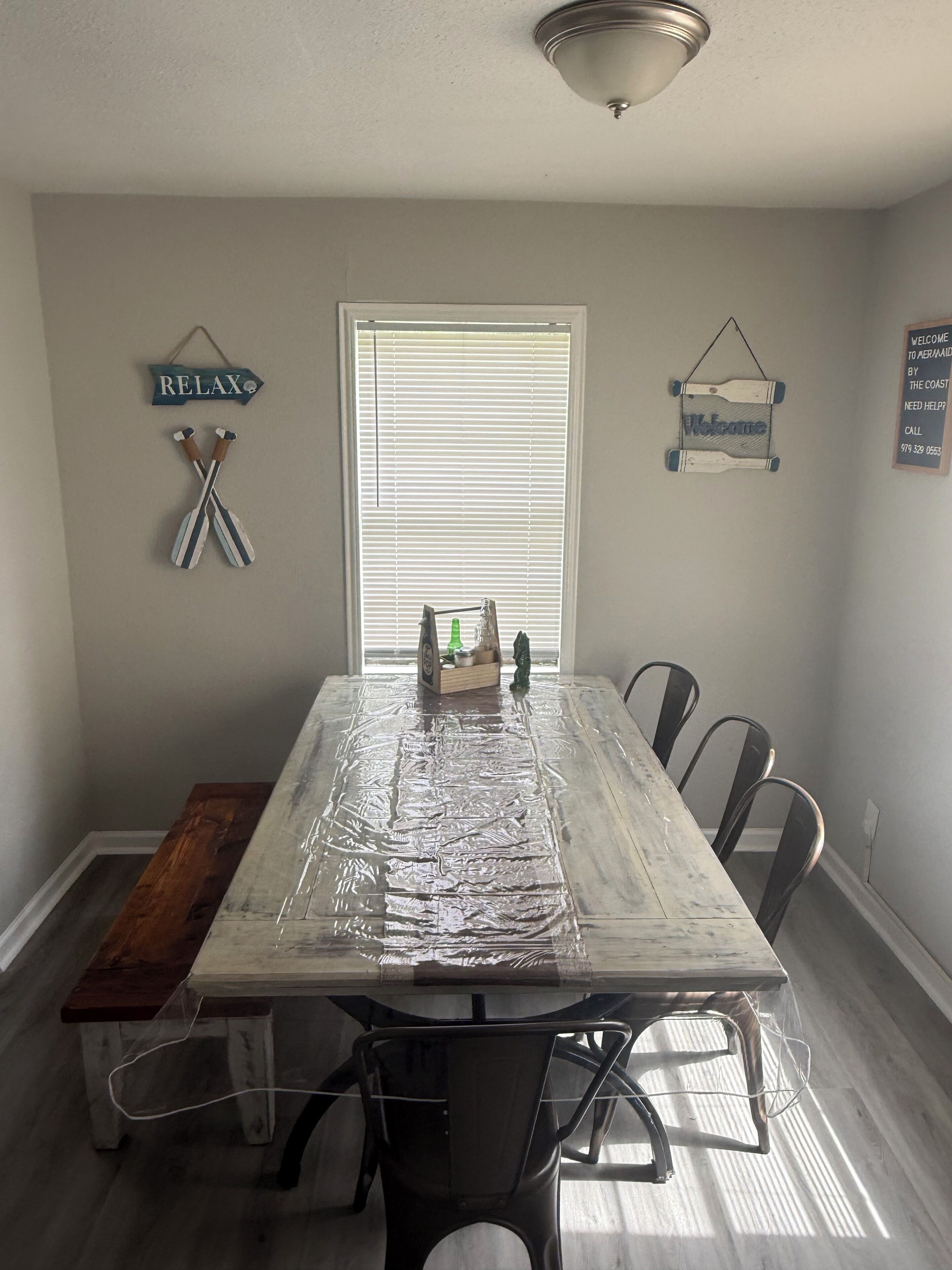 A dining room with a table covered in plastic, chairs, a wooden bench, and wall decorations including signs and a welcome sign.