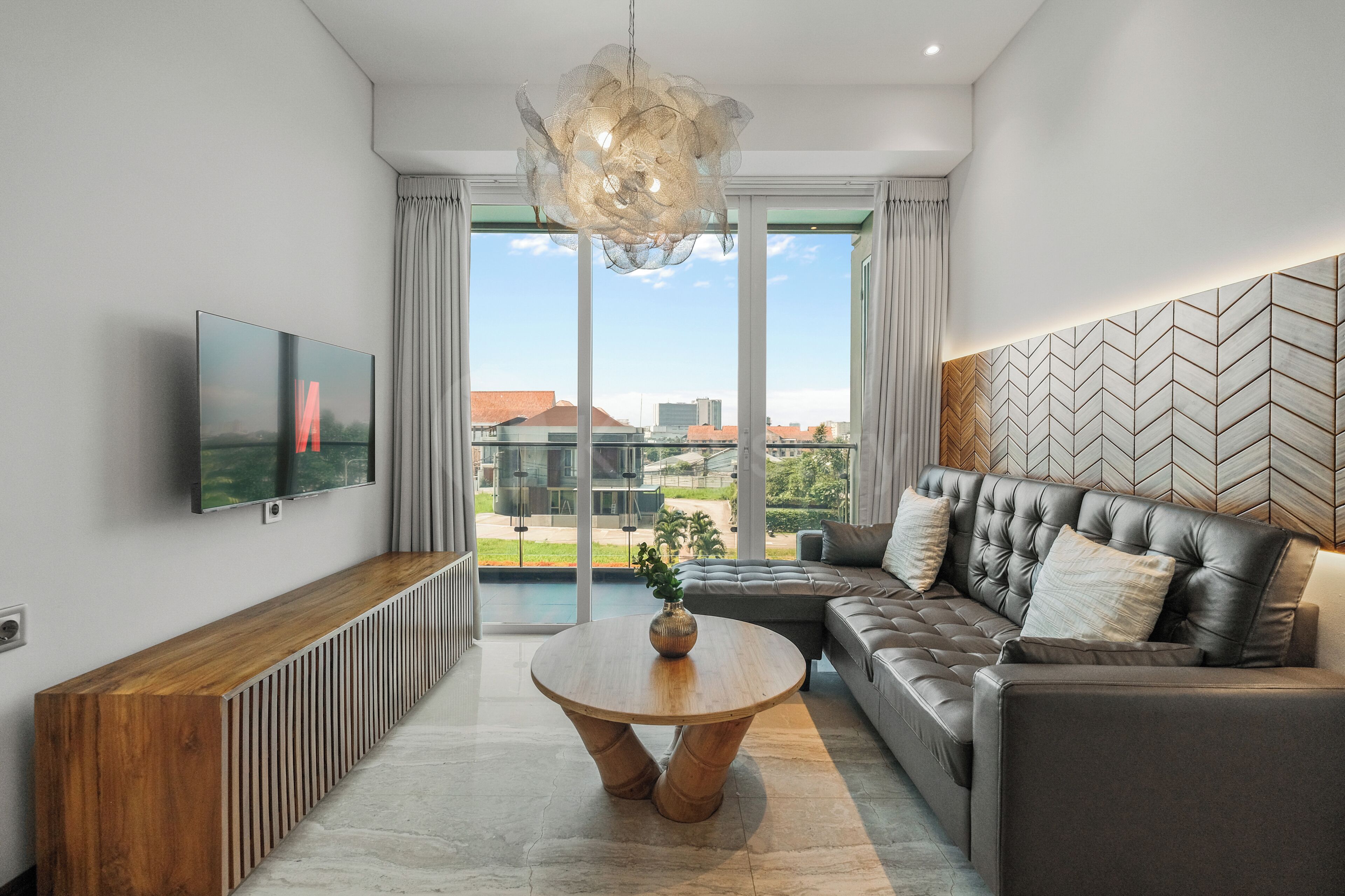 A modern living room with a large window, a sofa, a coffee table, and a TV mounted on the wall.
