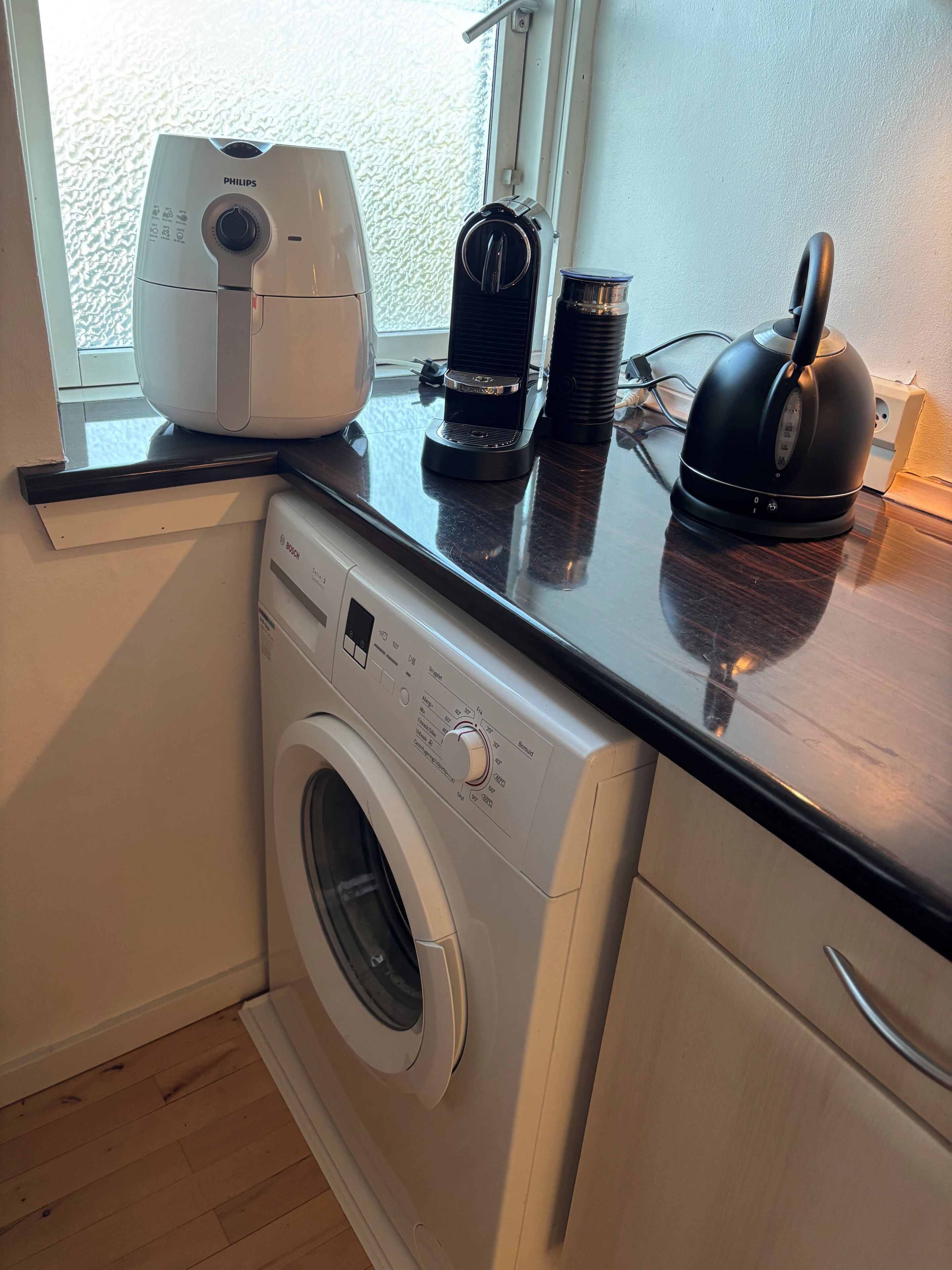 Une cuisine compacte équipée d’un lave-linge, d’une machine à café et d’une bouilloire posés sur le plan de travail.