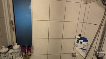 A bathroom with white tiles, a shower with a handheld showerhead, and a window with a blue tint.