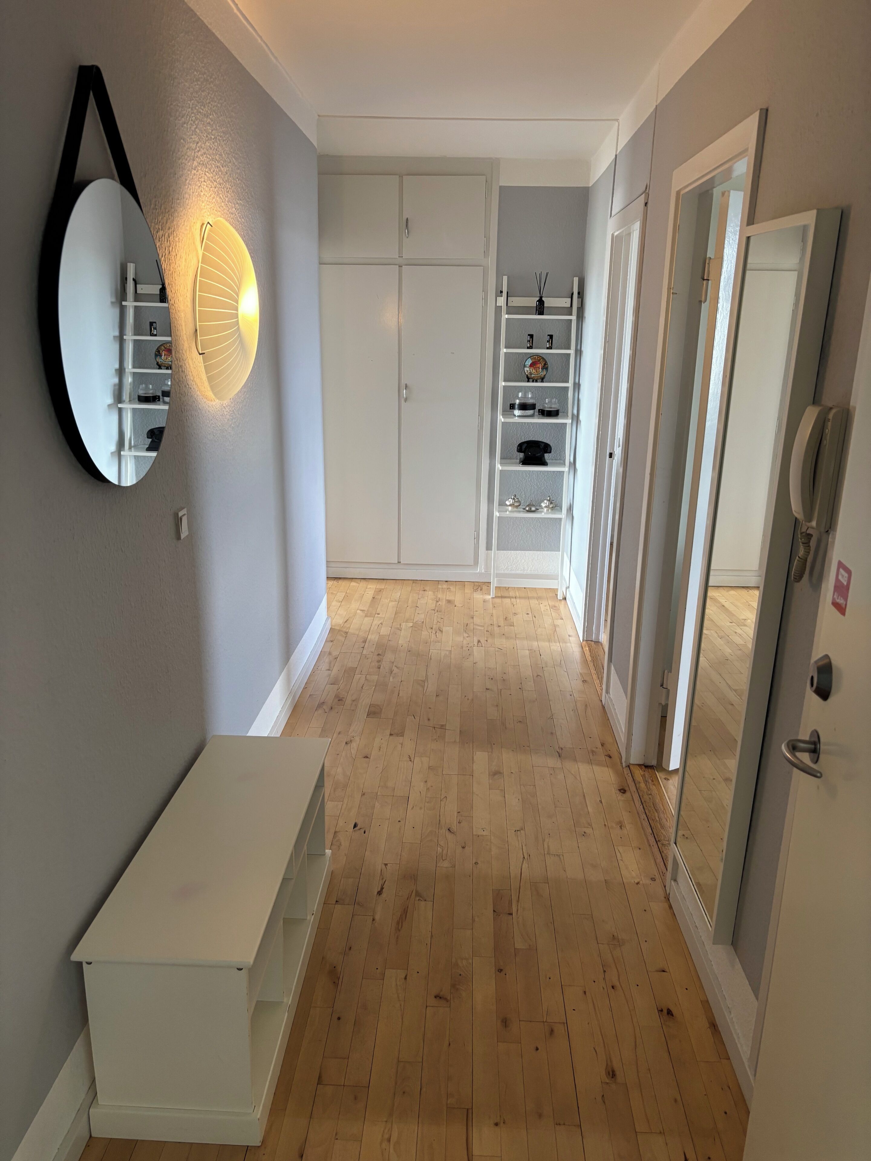 A hallway with a white cabinet, a mirror, and a shelf unit with various items.