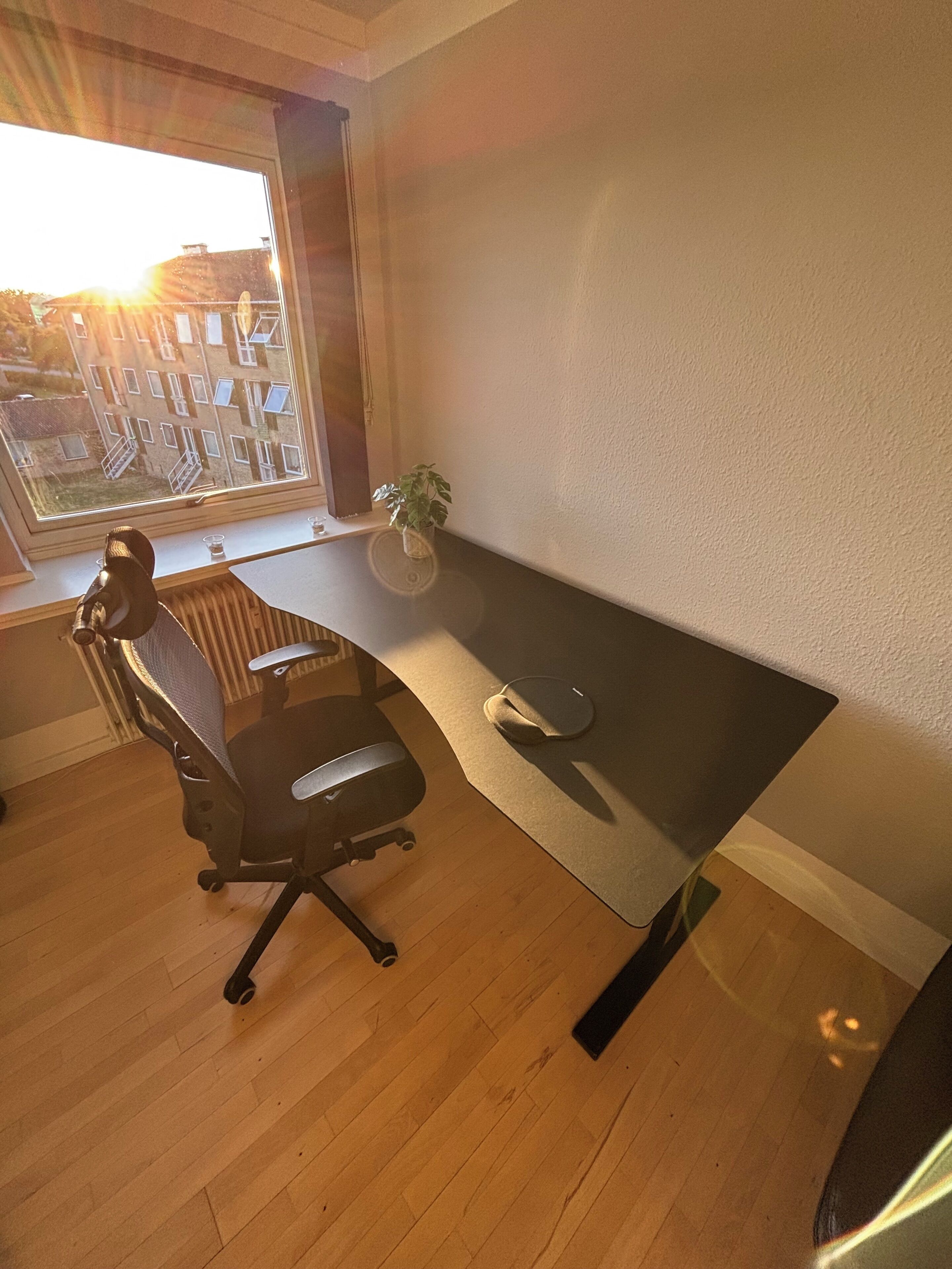 Ein Zimmer mit Holzboden, einem schwarzen Bürostuhl, einem kleinen Tisch, einer Topfpflanze und einem Fenster mit Blick auf Gebäude.