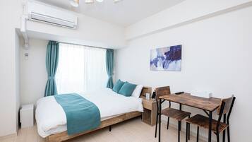 A bedroom with a bed, a desk, and chairs, an air conditioner, and a painting on the wall.