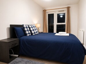 Ein ordentlich bezogenes Bett mit blauer Bettdecke und zwei Kissen, ein Nachttisch mit Lampe sowie ein Fenster mit Vorhängen.