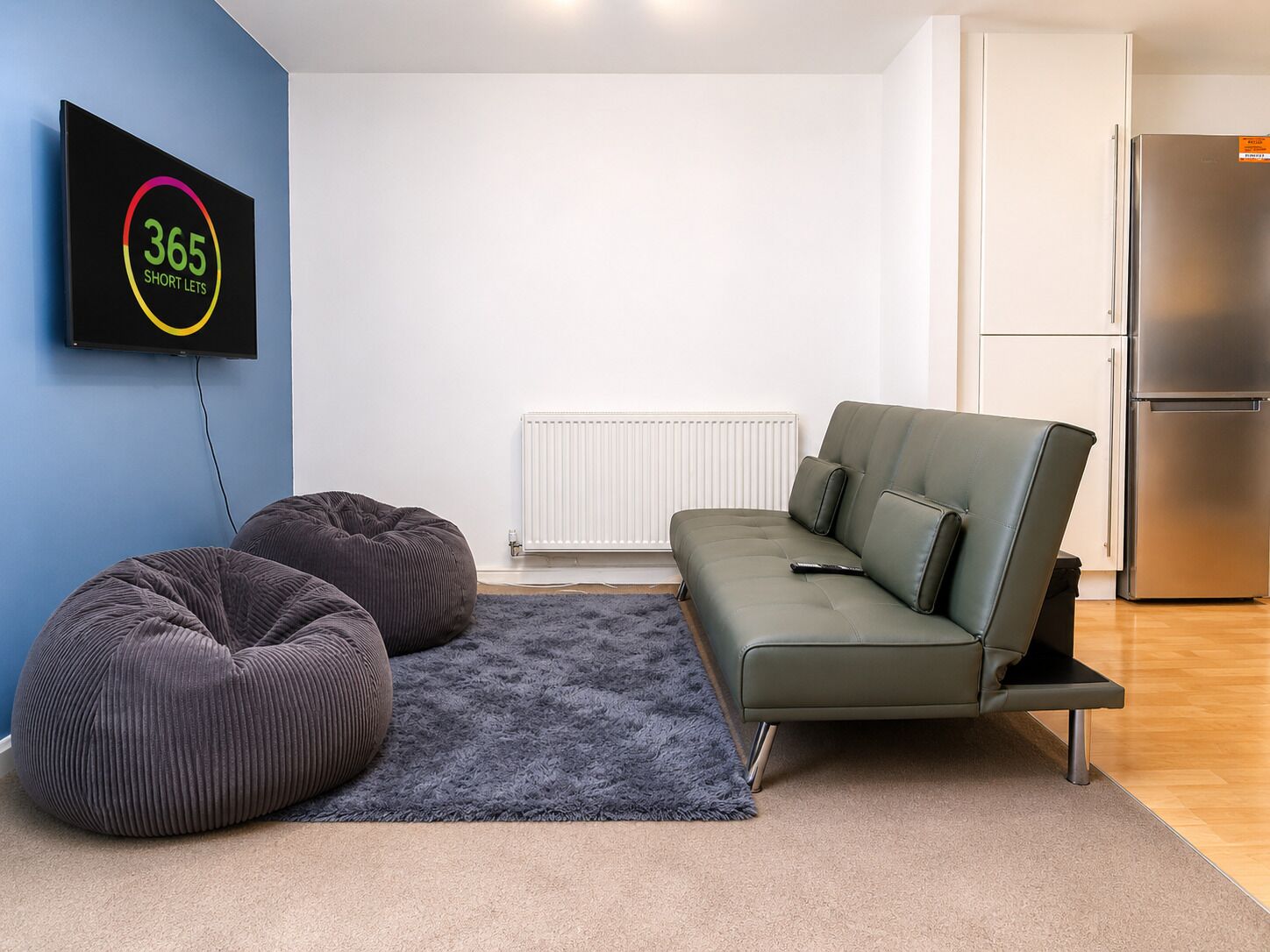 A modern living room with a sofa, two bean bags, a flat-screen TV, and a refrigerator.