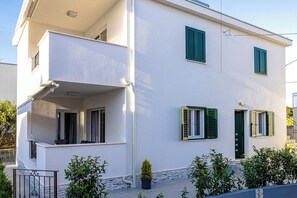Ein zweistöckiges Gebäude mit einem Balkon im oberen Geschoss, einer Tür mit grünem Rollo und einem kleinen Blumentopf im Vordergrund.