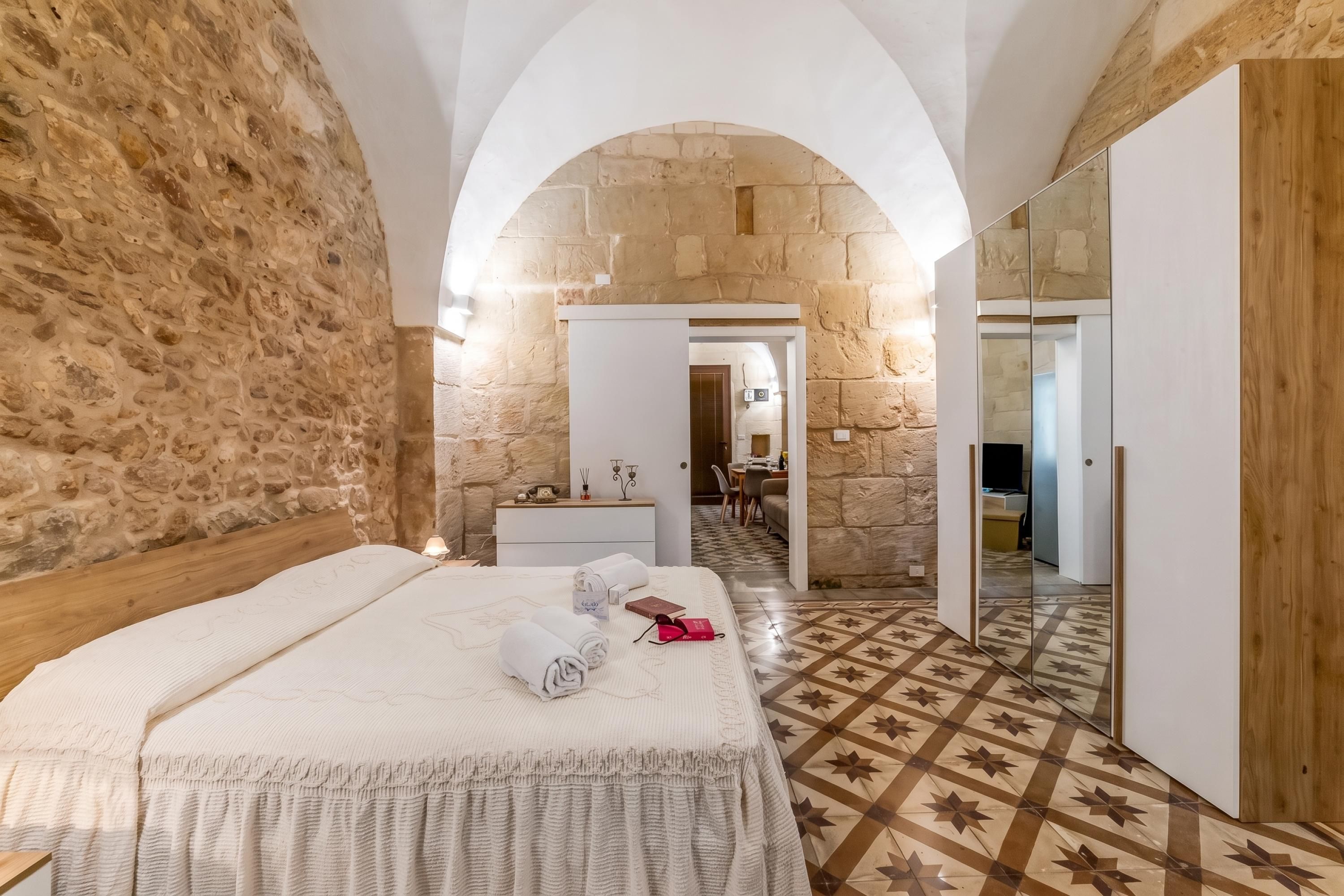 A hotel room with a stone wall, a wooden bed, a mirror, and a patterned floor.