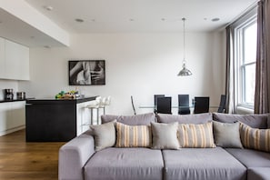 Ein modernes Wohnzimmer mit einem grauen Sofa, gestreiften Kissen, einer Küchenzeile mit weißen Schränken und einem Esstisch mit Stühlen.