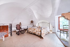 Ein Schlafzimmer mit einem Bett, einem Schaukelstuhl, einem Holzstuhl und einem Fenster mit Blick auf Grünfläche.