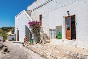 Un edificio blanco con puerta de madera, una escalera y un pequeño jardín con flores moradas.
