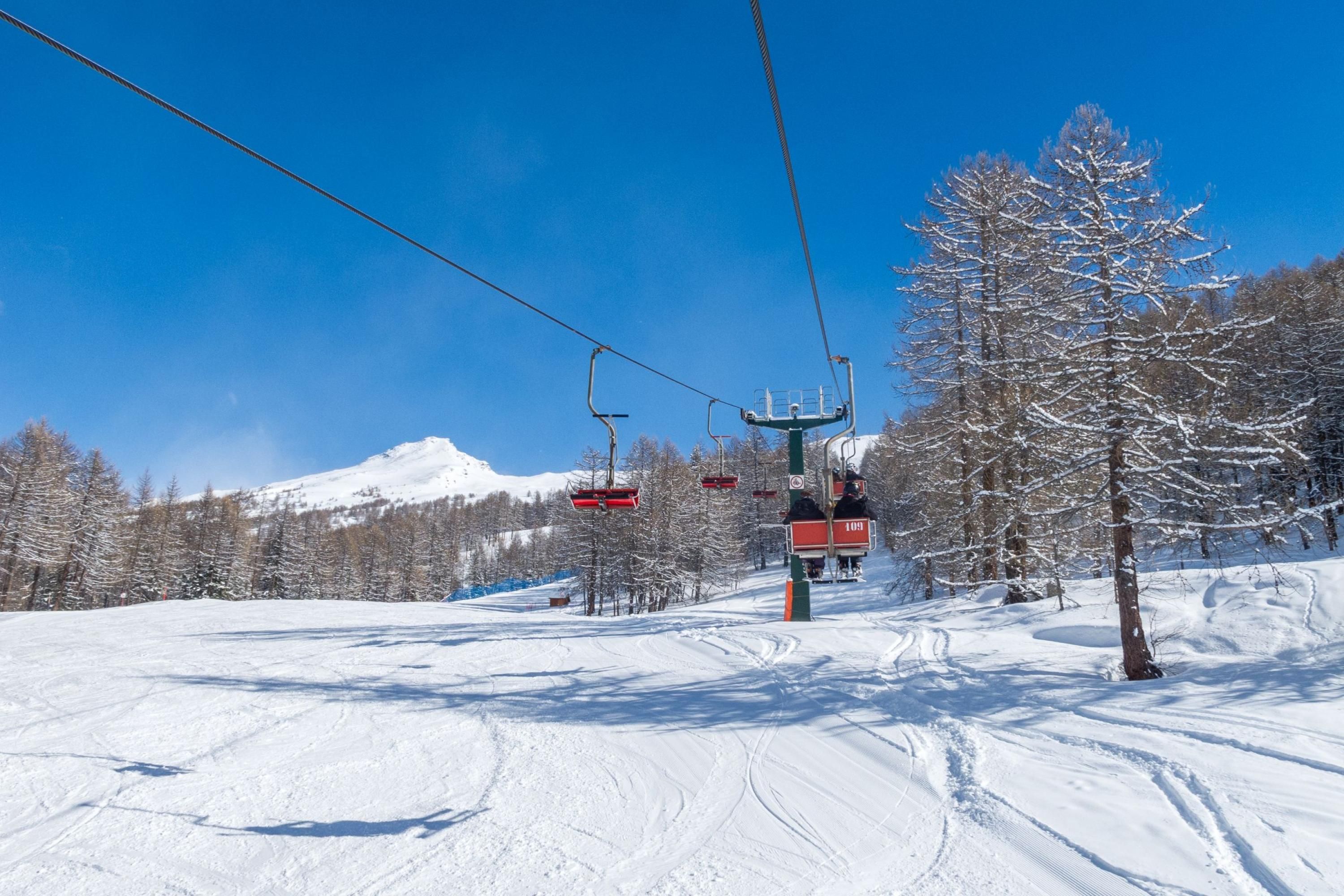 A ski lift with chairs moving along a snowy slope, surrounded by snow-covered trees and a clear blue sky.