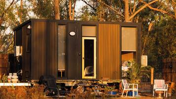 A small, dark-colored cabin with a single door and windows, surrounded by trees.