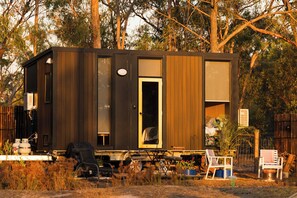 A small, dark-colored cabin with a single door and windows, surrounded by trees.