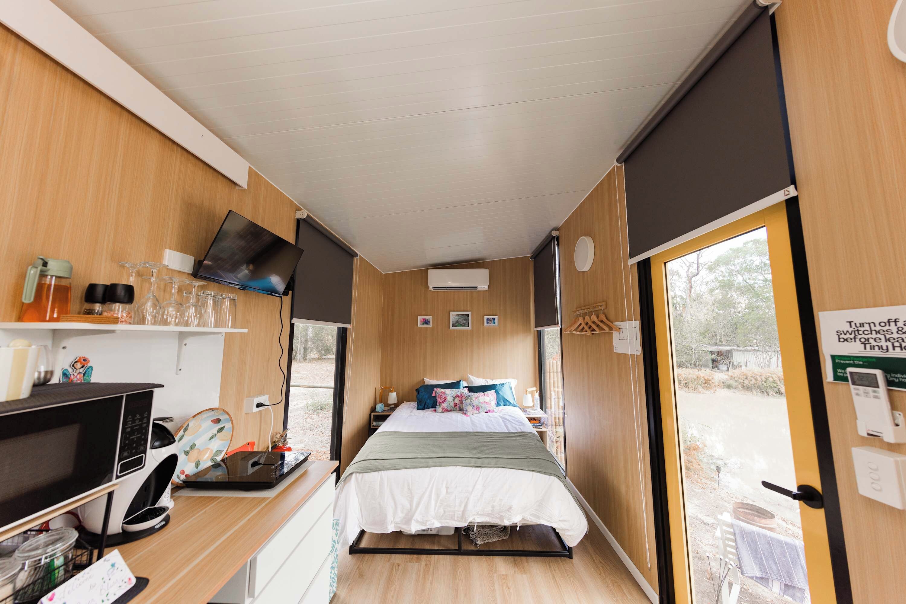 A compact room with a bed, a desk with a computer, a TV mounted on the wall, and a window providing a view of the outdoors.