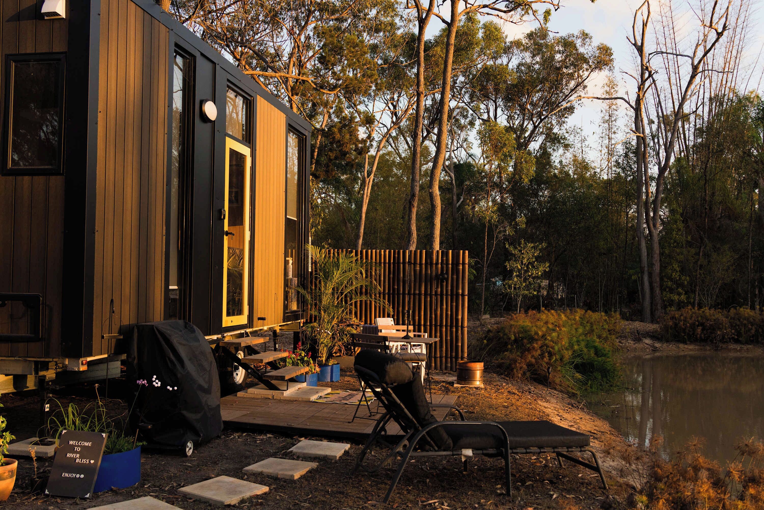 A wooden cabin with a deck, a grill, and a chair by a lake.