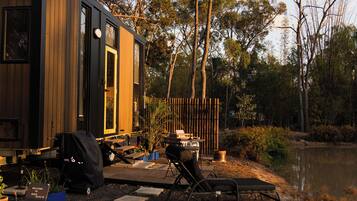 A wooden cabin with a deck, a grill, and a chair by a lake.