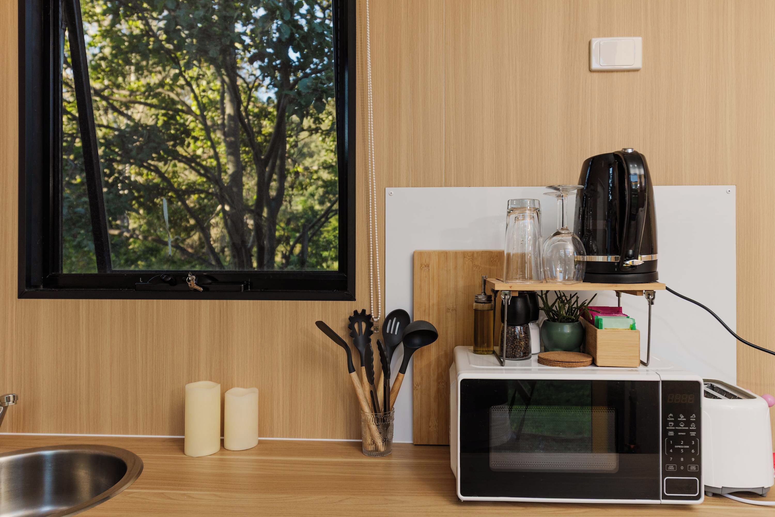 A kitchen with a microwave, toaster, and coffee maker on a wooden countertop, a window with a view of trees, and a sink.