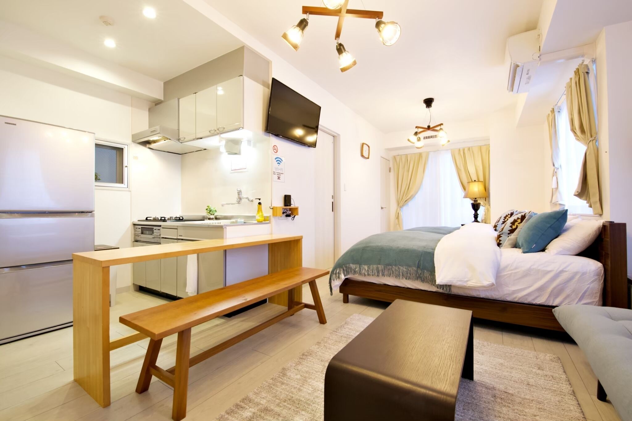 Una habitación de hotel compacta con kitchenette, una cama con manta verde, un banco de madera y una mesita de noche.
