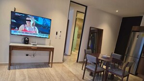 Uma sala de estar moderna com televisão de tela plana na parede, uma escrivaninha de madeira, uma mesa de jantar com cadeiras e um pequeno banquinho.